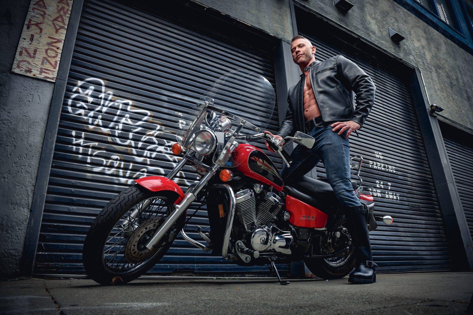 A man in a leather jacket astride a red motorcycle in a San Francisco alley of graffitied roll-up doors, shot from a low heroic angle.