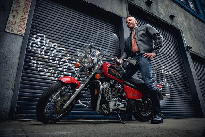A man in a leather jacket astride a red motorcycle in a San Francisco alley of graffitied roll-up doors, shot from a low heroic angle.