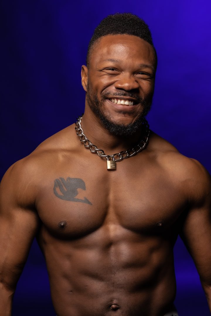 A Black man with a chain-and-lock collar breaks into a wide, easy smile on a saturated electric-blue studio ground.
