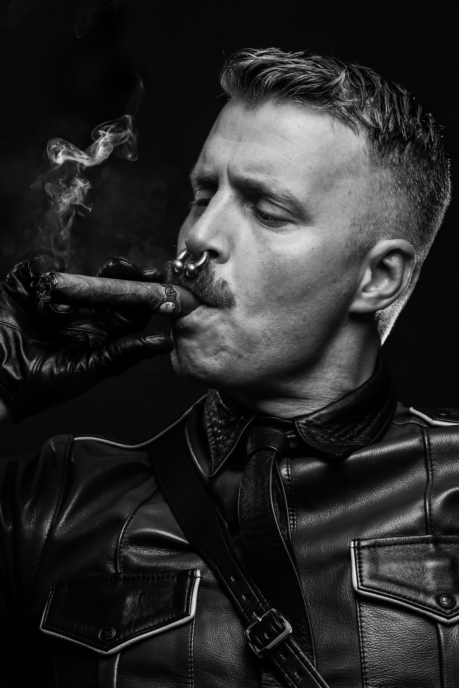 A high-contrast black-and-white portrait of a man in leather with a cigar, deep shadow holding the frame.