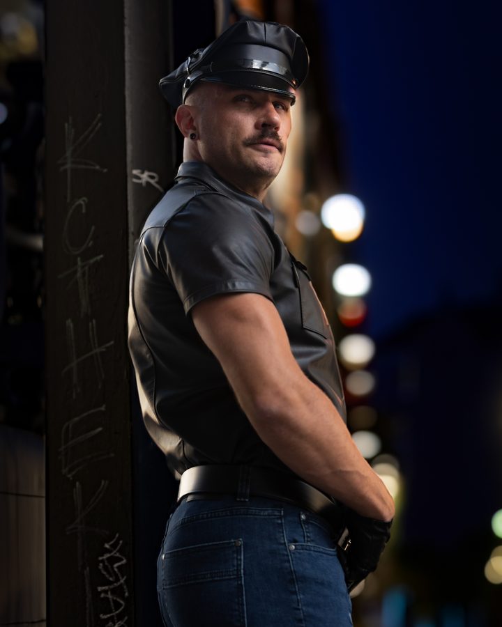 A man in leather turning three-quarters away on a night street, city lights softened to bokeh behind him.