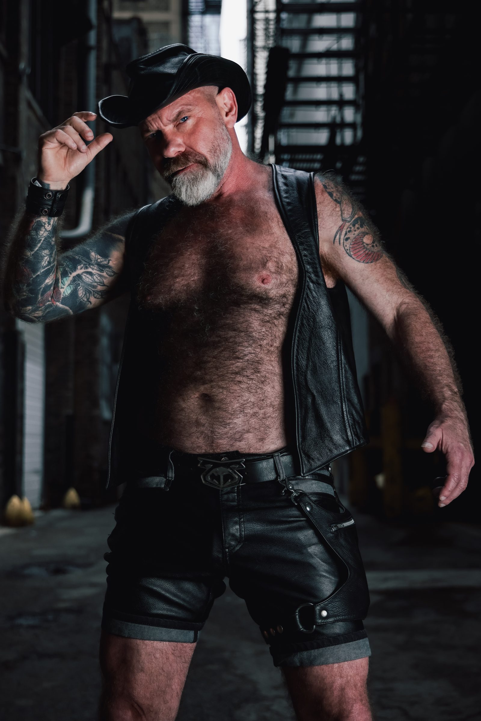 A silver-bearded man in a leather vest, cap, and shorts tips his cap brim in a dim San Francisco alley, a fire escape rising behind him.