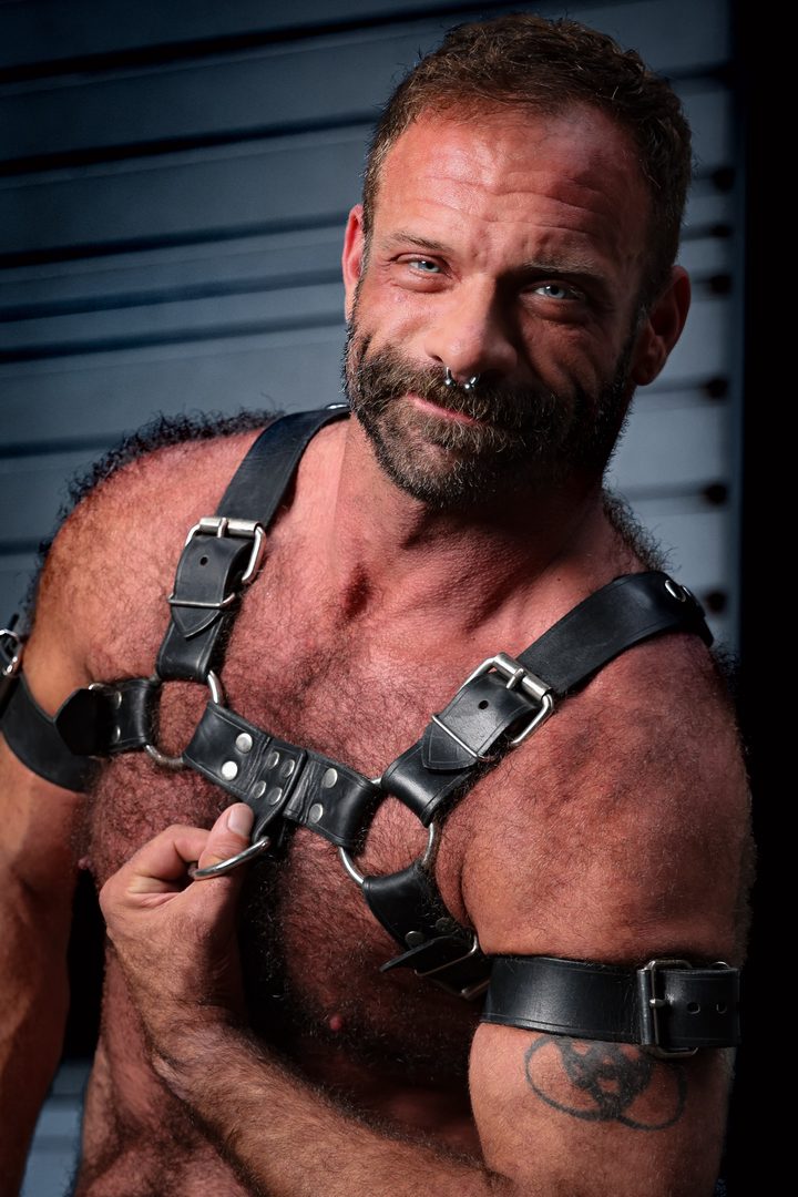 A silver-bearded, broad-chested man in a leather harness gives the lens a warm half-smile, lit hard against a deep-blue garage door.