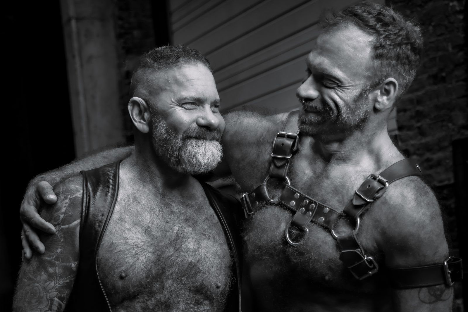 A black-and-white frame of two leather men sharing a laugh, one resting his head toward the other's shoulder in an easy, warm moment.