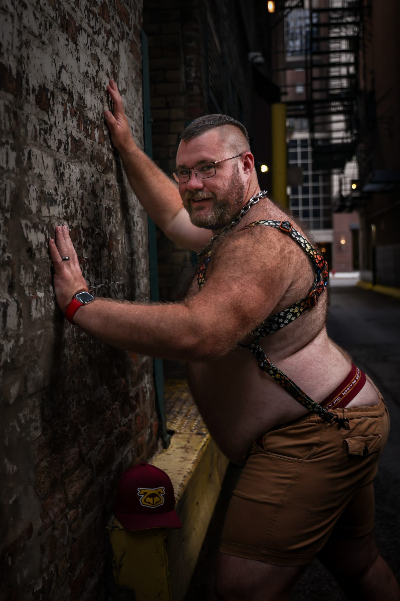 A broader-bodied, hairy-chested man in glasses and a Pride-trimmed harness leans against a brick alley wall, glancing back with an easy grin.