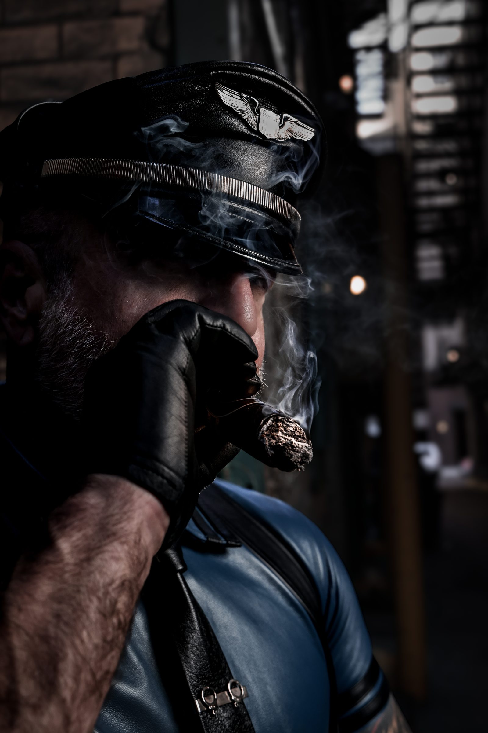 A profile of a man in a winged-insignia Muster cap drawing a cigar with a gloved hand, smoke threading the lamplit alley behind him.