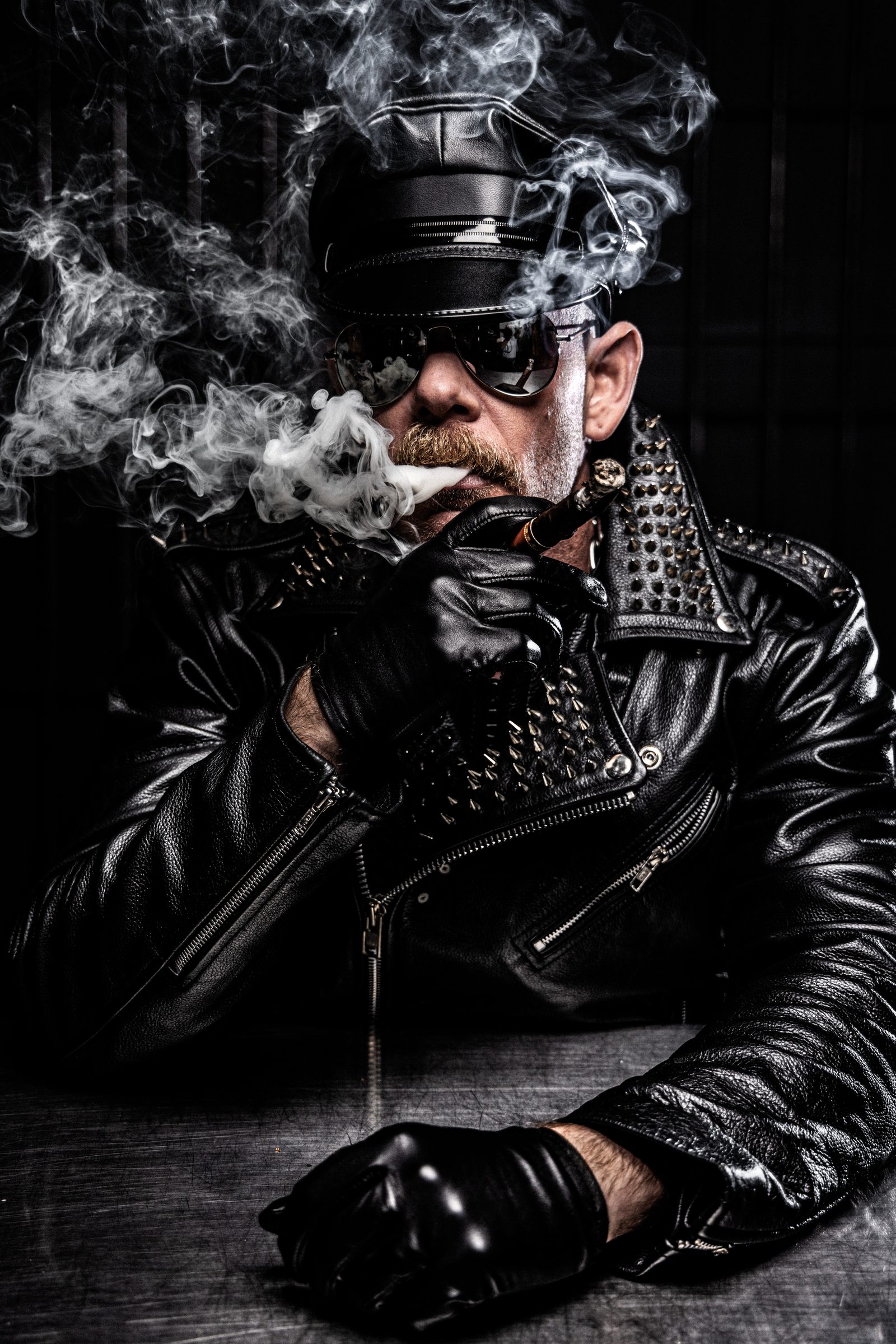 A man in a leather Muster cap with a cigar, lit by hard studio strobe through billowing smoke against a near-black ground.