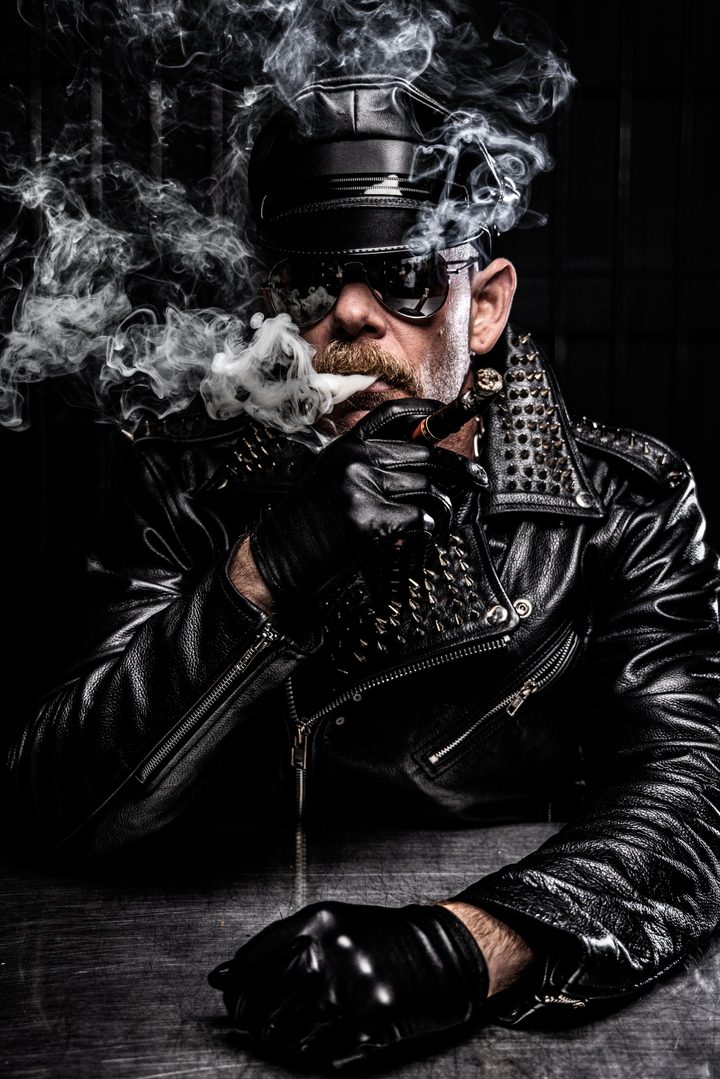 A man in a leather Muster cap with a cigar, lit by hard studio strobe through billowing smoke against a near-black ground.