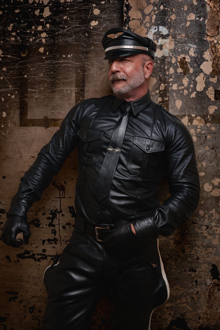 A black-and-white portrait of a silver-bearded man in a full leather uniform and Muster cap, low-key light raking across him on a distressed wall.