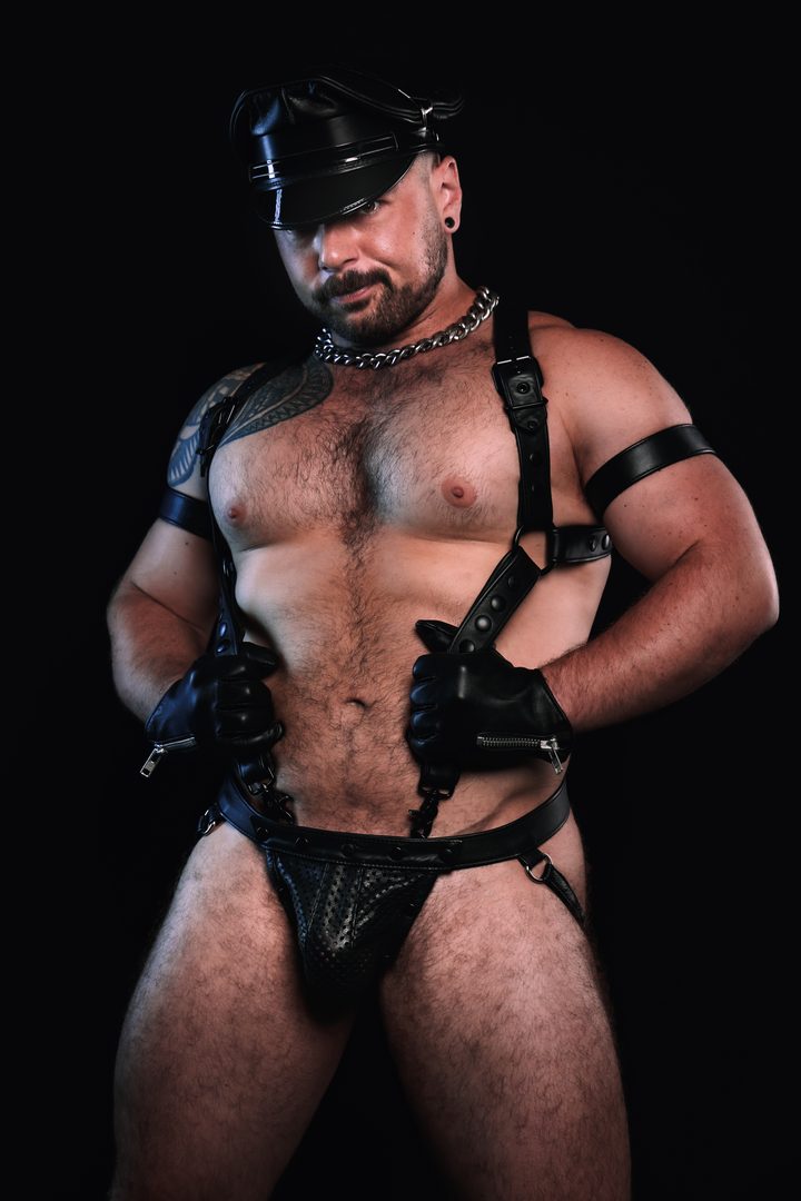 A bearded man in a leather Muster cap, harness, and gloves stands square against near-black, hands resting at his harness in classic leather grammar.