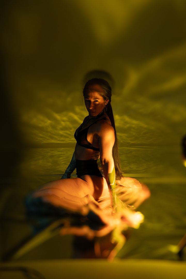 A seated figure lit toxic green and gold, hands raised, the gelled light shaping the form against a dark ground.