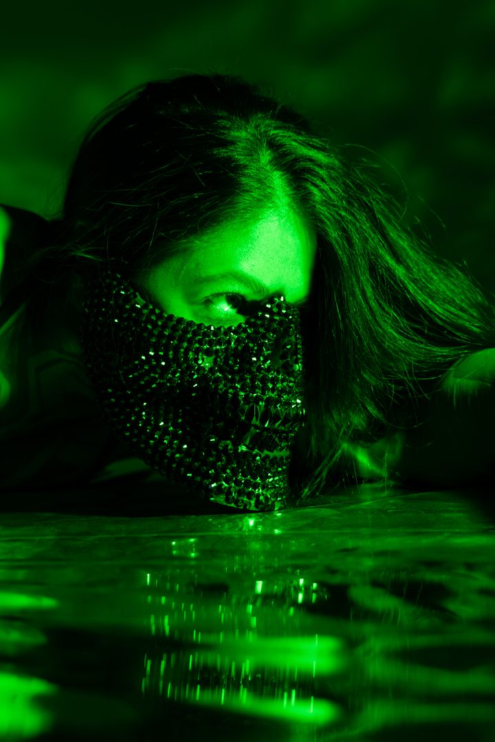 A figure in a beaded mask lit toxic green, low to a reflective floor that mirrors the light back in rippling streaks.