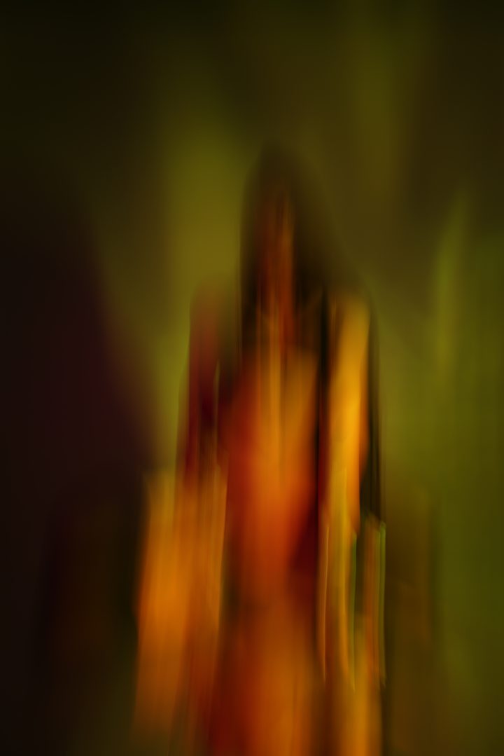 A standing figure pulled into vertical streaks of amber, red, and olive by long exposure, almost wholly abstract.