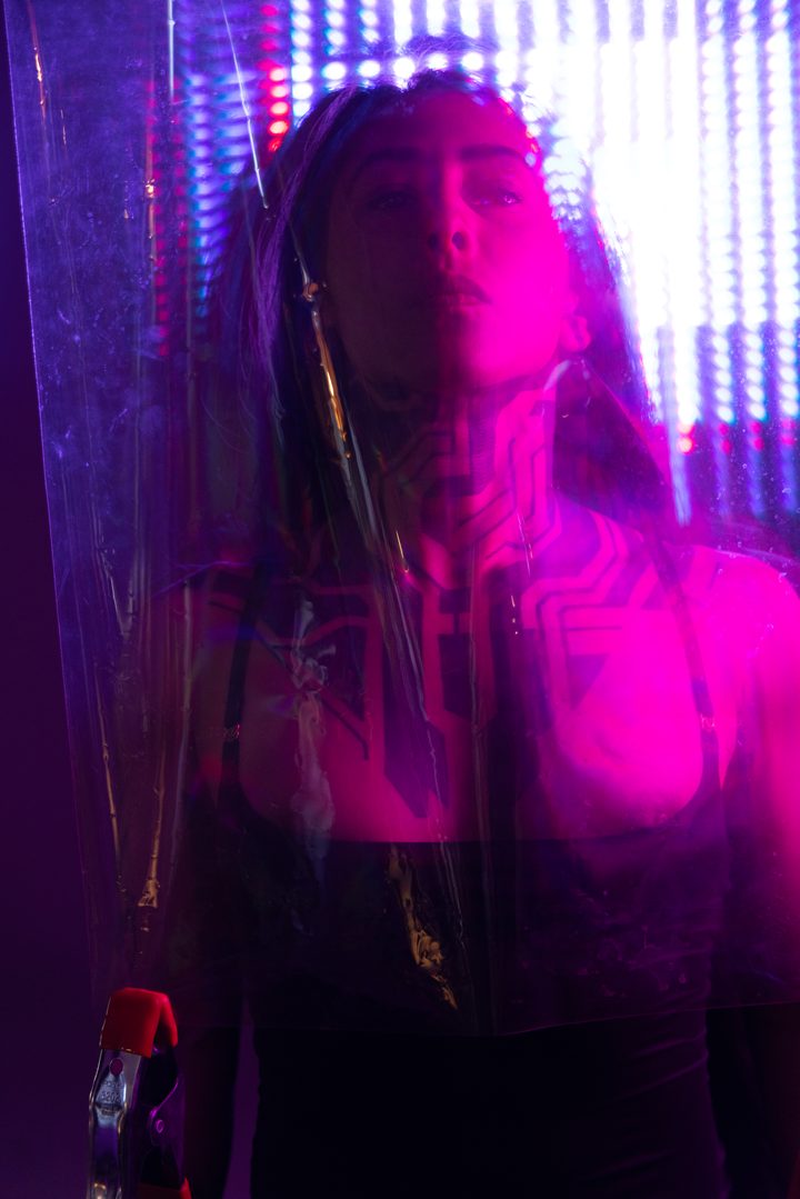 A figure behind a sheet of plastic in magenta and blue club light, projected linework doubling across the body.
