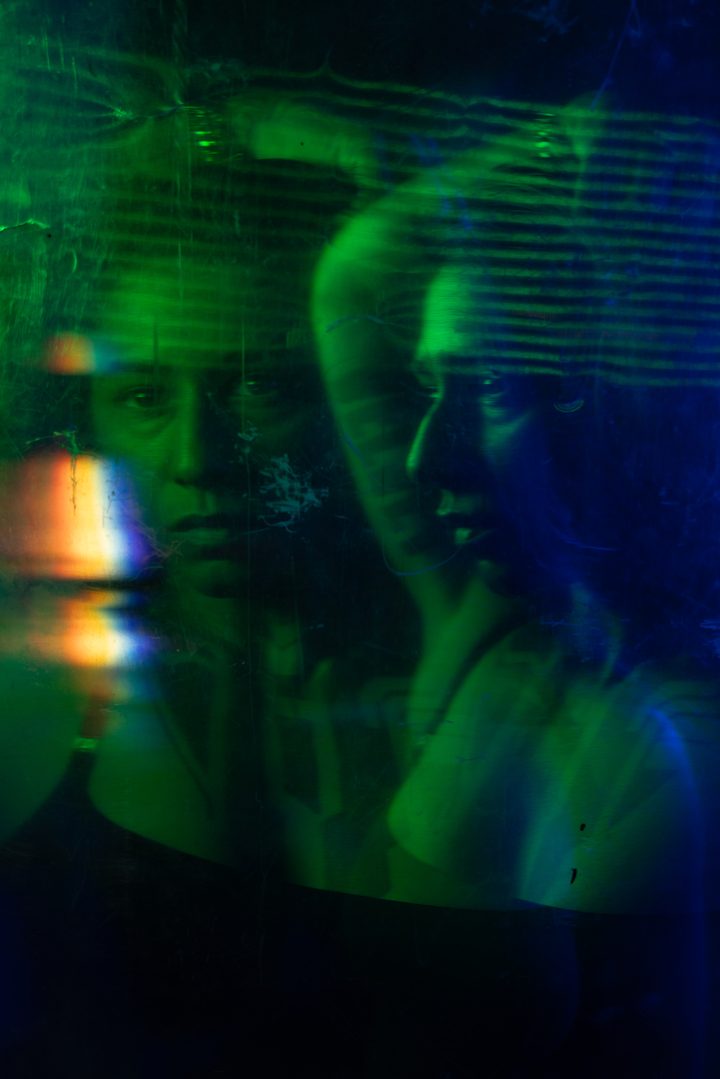 A face seen through a sheet of scratched, wet acetate in violet and white light, the surface fracturing the portrait.
