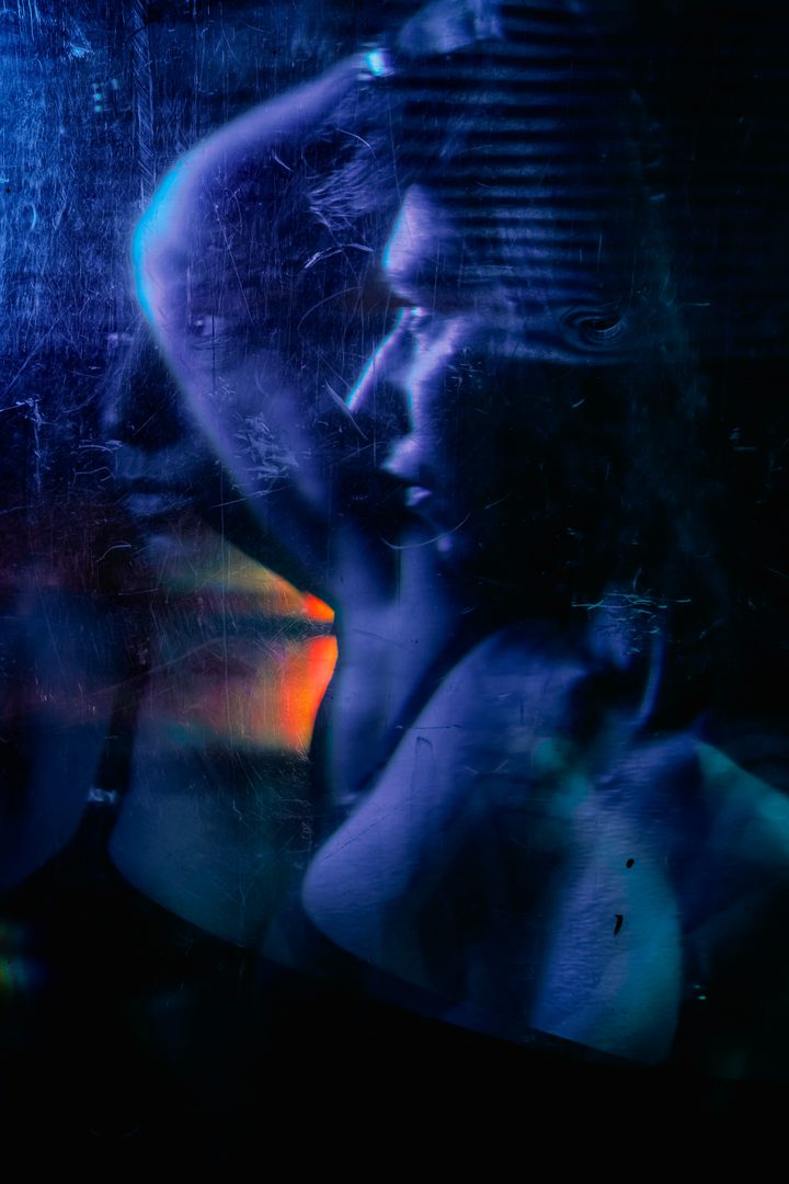 A figure behind textured glass, reaching toward the surface, the gesture broken into soft cool highlights by the projection.
