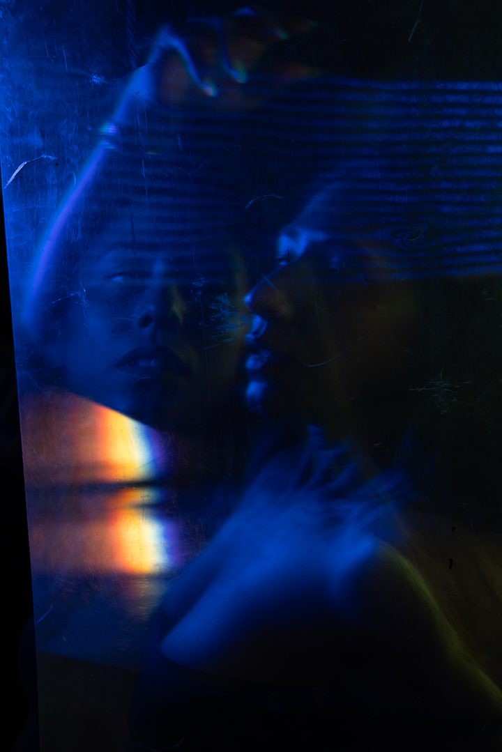 A reclining figure under green projected light, hands raised, the body lit low against a dark, textured ground.